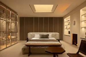 Modern minimalist bedroom with a bouclé upholstered bed, wooden bench at the foot, glass-enclosed walk-in closet on the left, and built-in shelving with fireplace on the right.