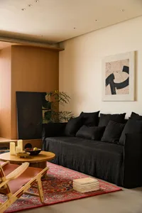 Modern minimalist living room featuring a black linen sofa, round oak coffee table, wooden lounge chair with red leather seat, Persian rug, built-in oak cabinetry, and abstract wall art.