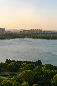 Aerial view at sunset showing a wide lake surrounded by dense green trees in the foreground and a modern city skyline with numerous high-rise buildings in the background under a golden sky.
