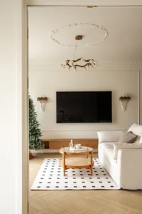 View into a bright living room with white sofa, round oak coffee table on a black and white checkered rug, wall-mounted TV, and a decorated Christmas tree.
