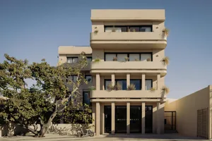 Front view of a multi-story beige concrete residential building with curved balconies, large windows, and potted plants under a clear blue sky.