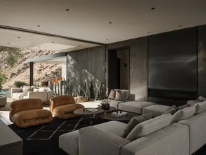 Modern desert living room featuring concrete walls, large grey sectional sofa, two cognac leather lounge chairs, round coffee tables on a dark rug, with desert landscape and cacti visible through open sliding doors.