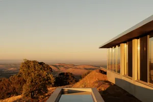 Modern concrete and glass villa with large windows and an infinity pool overlooking a vast golden landscape at sunset.