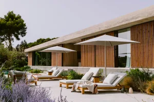 Modern concrete villa with vertical timber cladding, featuring multiple teak lounge chairs with white cushions and large white umbrellas on a spacious concrete terrace surrounded by lavender and Mediterranean landscaping.