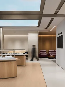 Modern luxury jewelry boutique interior with curved oak display counters, beige sofas, burgundy leather armchairs, wooden slat accent wall, and large skylight.