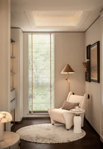 Cozy reading nook in a modern apartment featuring a cream boucle armchair, brass floor lamp, round patterned rug, and large window overlooking green trees.
