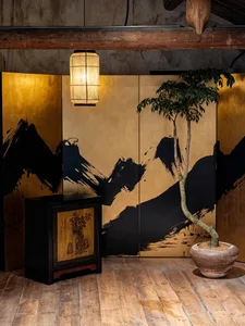 Japanese-style folding screen with black ink mountain landscape on gold leaf panels, accompanied by a black wooden cabinet, hanging paper lantern, and a tall bonsai tree in a ceramic pot on wooden flooring.