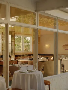 Elegant coastal restaurant interior featuring round tables with white tablecloths, oak banquette seating, large multi-pane windows overlooking greenery, and warm natural light.
