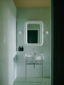 Narrow powder room with light mint-green ceramic tile walls and floor, white ceramic wall-mounted sink with chrome legs, oversized white chunky pipe-style mirror, and two black soap dispensers.