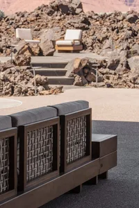 Modern dark teak outdoor sofa with grey cushions and rope detailing positioned on a stone terrace overlooking rugged desert rocks and two background lounge chairs.