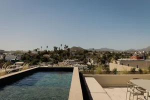 Modern rooftop terrace with a rectangular infinity pool, concrete deck, outdoor lounge seating, and a panoramic view of a desert city and distant mountains under a clear blue sky.