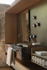 Luxury bathroom featuring a thick marble vanity with integrated sinks, brushed nickel faucet, olive green wall, and rough stone feature wall with floating wooden shelves holding rolled towels.