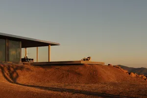 Modern minimalist house with large glass windows and concrete terrace overlooking a golden desert hill at sunset, with mountains in the distance.