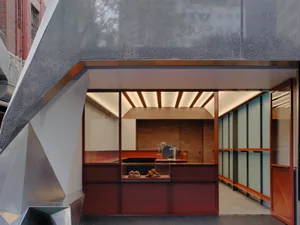 Modern café entrance with angular metallic facade and interior walnut service counter displaying pastries under warm linear lighting.