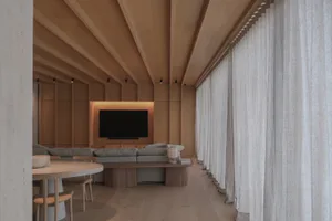 Modern minimalist living room featuring a dramatic sloped oak ceiling, built-in entertainment wall with backlit TV, light grey sectional sofa, round marble dining table, and floor-to-ceiling sheer curtains.