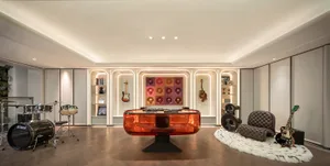 Modern music room featuring a transparent orange acrylic DJ console, black drum kit, electric guitars, built-in illuminated shelving, and a brown tufted lounge chair on a white fur rug.