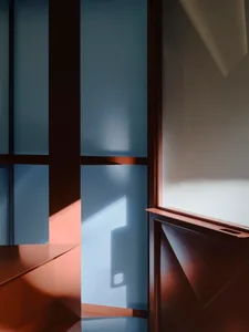 深色胡桃木板材與層架的特寫建築細節，強烈的晨光在牆面上投射出藍色與暖橘色光影。