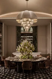 Luxurious formal dining room featuring a large round black marble table, velvet dining chairs, dramatic floral centerpiece, and a massive contemporary fabric chandelier against a dark green marble accent wall.
