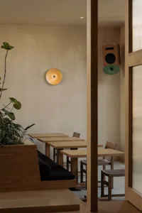Minimalist cafe dining area featuring solid oak tables and chairs, a built-in wooden banquette with black cushions, indoor plants, a modern wall sconce, and a hanging wooden speaker with green cone.