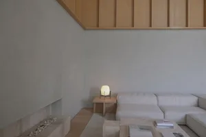 Modern minimalist living room featuring a light oak slat ceiling, concrete walls, a modular off-white sofa, travertine coffee tables, and a glowing table lamp.