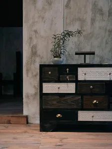 Vintage-style black reclaimed wood sideboard with white carved drawers and brass pulls, placed against a textured concrete wall with a potted plant on top.