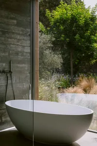 Modern minimalist bathroom featuring a white freestanding oval bathtub against a raw concrete wall, with large glass windows overlooking a lush green garden with trees and wildflowers.