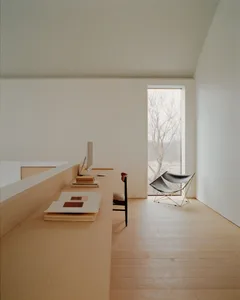 Minimalist home office featuring a long oak built-in desk, computer monitor, wooden chair, and two black butterfly chairs by a large window with natural light and bare trees outside.