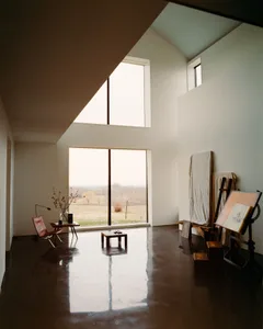 寬敞的極簡藝術工作室，擁有高挑天花板、磨光混凝土地板、皮革蝶形椅、擺放乾燥樹枝的木製邊桌，以及右側的大型畫架與畫布。