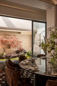 Modern luxury dining room featuring a dark marble oval table, velvet patterned dining chairs, and a view of a Japanese-style garden with red maple tree and large rocks.
