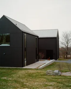 Modern black barn house with vertical wood cladding, large windows, metal roof, concrete terrace and gravel path set in a rural landscape.