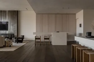 Modern minimalist open-plan living, dining and kitchen with concrete island, light oak cabinetry, dark oak flooring and sculptural wooden stools.
