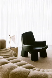 Modern living room corner featuring a black sculptural armchair, a beige boucle sofa, a white table lamp on a fluted side table, and floor-to-ceiling sheer white curtains.