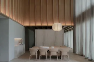 Modern minimalist dining room featuring a long oak dining table with beige upholstered chairs, a large ribbed pendant lamp, vertical wood slat ceiling, and a prominent concrete accent wall.