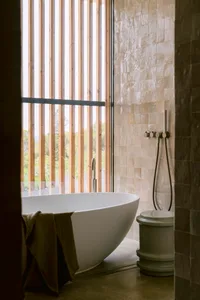 Modern minimalist bathroom featuring a white freestanding oval bathtub, beige travertine tiled walls, vertical wooden slats with mountain view, and a matte grey vessel toilet.