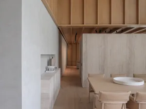 Modern minimalist dining room featuring travertine walls, a light oak dining table with beige leather chairs, built-in stone console, and warm wooden slat ceiling with track lighting.