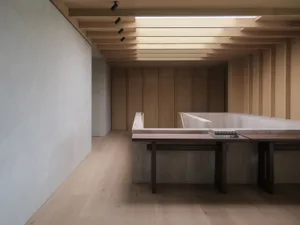 Modern minimalist interior featuring a concrete reading nook with a dark oak bench and stacked books, under a sculptural wooden ceiling with recessed lighting.