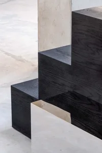 Close-up view of a modern staircase featuring thick black oak wooden treads with visible grain, cantilevered over a light gray polished concrete floor, with a textured concrete wall in the background.