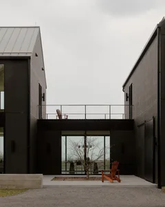 Modern black charred wood house exterior featuring a central courtyard with large glass windows, a small tree, and two wooden Adirondack chairs on an upper balcony and ground level.
