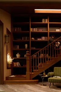 Warmly lit interior featuring a walnut staircase with integrated bookshelves, a green striped armchair, and a person browsing books on the upper level.