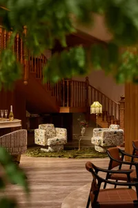 Interior view of a luxurious lounge area featuring two floral-patterned armchairs, a green velvet rug, wooden staircase with turned balusters, and warm ambient lighting from a fringed floor lamp.