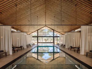 Long indoor lap pool in a serene spa with high vaulted wooden ceiling, white curtains, and wooden lounge chairs on both sides.