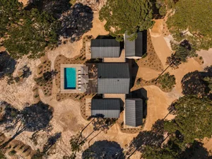 Aerial view of a modern desert villa featuring dark metal roofs, a rectangular turquoise swimming pool, wooden decks, and surrounded by arid landscape with scattered pine trees.