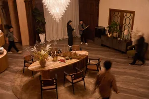 High-angle view of a spacious modern dining room with a large irregular travertine dining table, brown leather chairs, a large crystal chandelier, and people moving in the background.