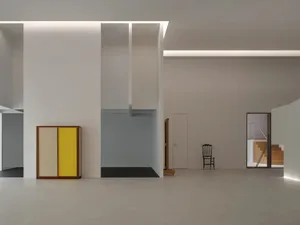 Modern minimalist entrance hall with high white walls, recessed ceiling lighting, a two-tone yellow and cream lacquered wood cabinet, and a black wooden chair in the distance.