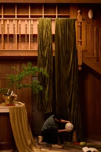 Man kneeling on the floor arranging a decorative gourd beside heavy olive-green velvet curtains hanging under a carved wooden staircase with turned balusters and warm wood paneling.
