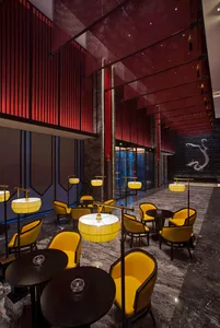 Luxurious hotel lobby lounge featuring bright yellow velvet armchairs, black round tables, dramatic red vertical slat walls, and grey marble flooring with a large white dragon mural in the background.