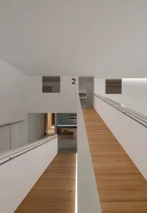 Modern double-height interior corridor featuring light oak wood flooring, white walls, continuous handrails, and linear LED lighting strips along the floor edge.