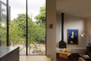 Contemporary living room featuring a tan leather armchair, black metal fireplace, large Mona Lisa painting, and floor-to-ceiling glass doors opening to a green garden.