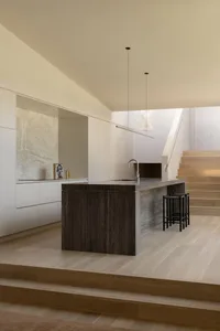 Modern minimalist kitchen featuring a dark marble island, white handleless cabinetry, light wood flooring, and a wooden floating staircase in the background.