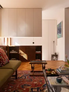 Modern minimalist living room featuring built-in beige upper cabinets, walnut lower storage with louvered doors, olive green sofa with patterned cushion, Persian rug on herringbone wood floor, and natural sunlight streaming in.